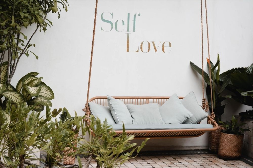 outdoor swing with plants and a self love sign above it